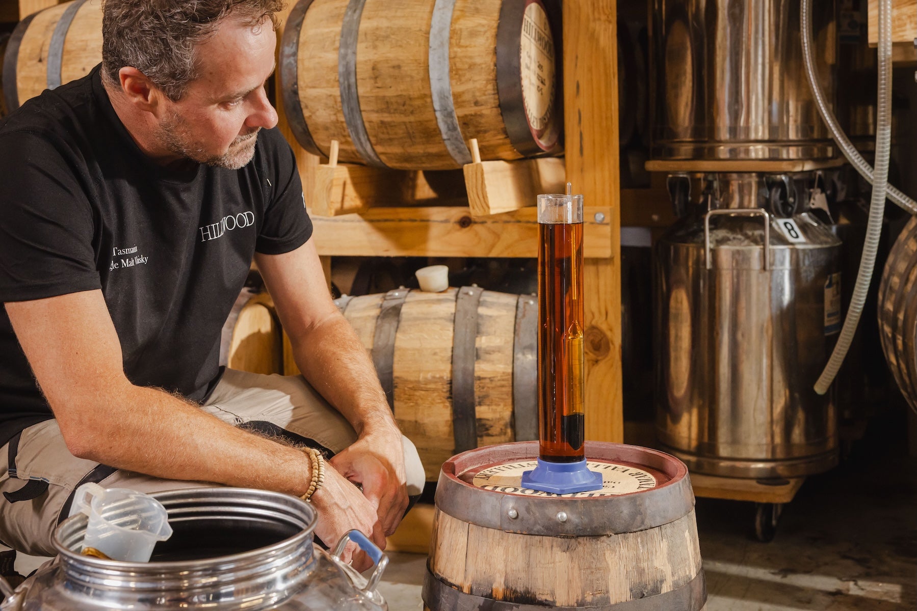 Ollie testing the Single Malt Whisky to determine its ABV. A measured about i in a measuring cylinder resting flat on a cask and Ollie is reading the temperature of the single malt whisky.